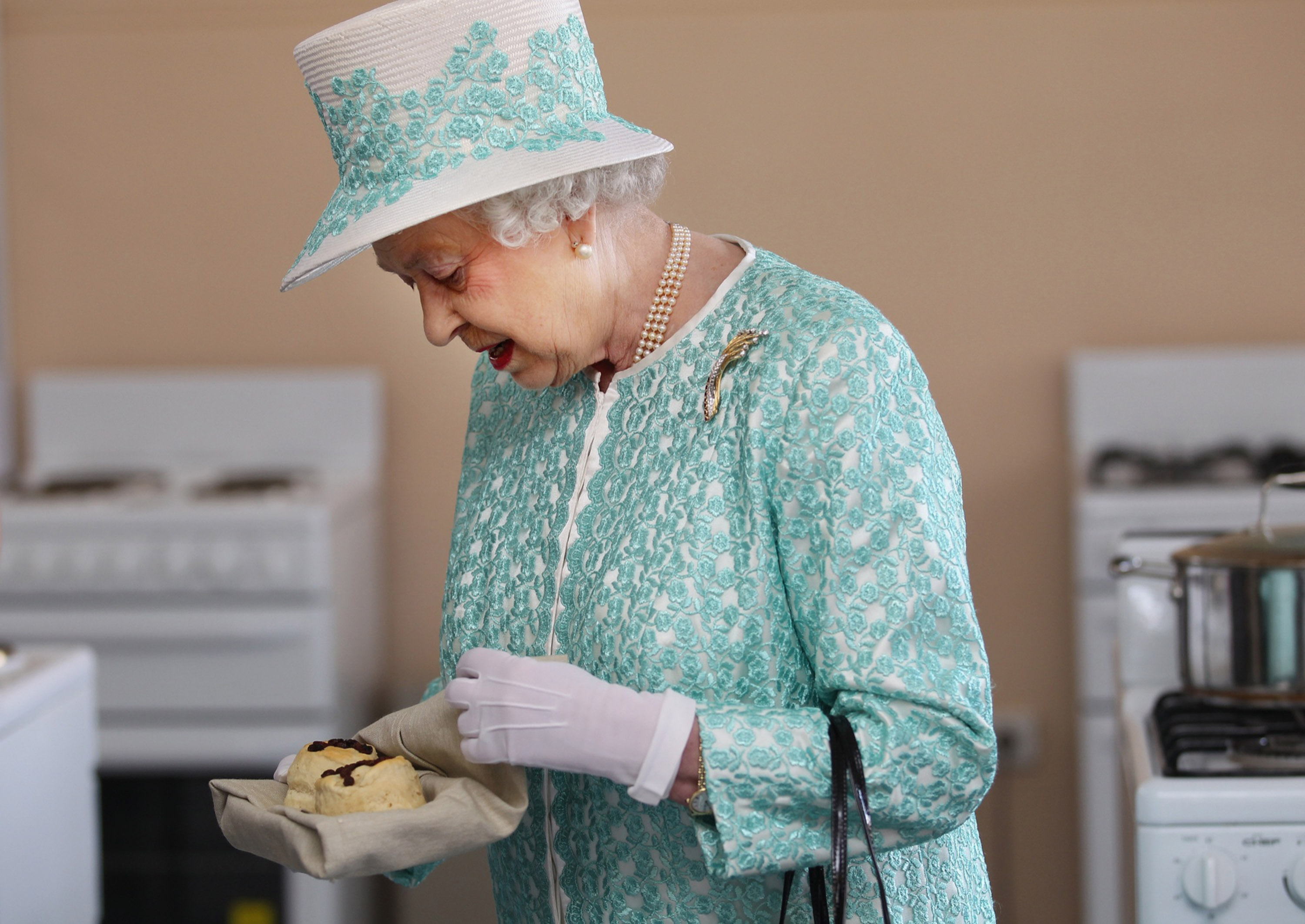 Queen Elizabeth wears a white and turquoise coat, a white hat adorned with embroidery, and white gloves, while eating a scone