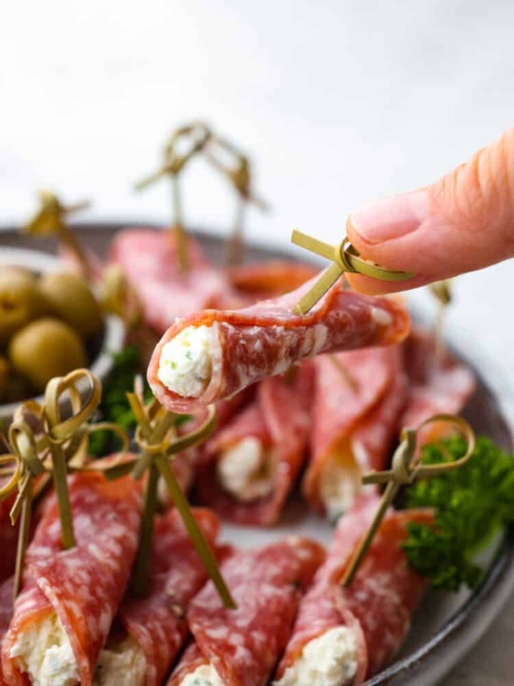 If you are looking for the perfect snack or appetizer, salami roll ups are your new best friend! Salami is wrapped around seasoned cream cheese for the perfect savory bite every time. A roll up being held up by a hand over a plate of other salami roll ups.