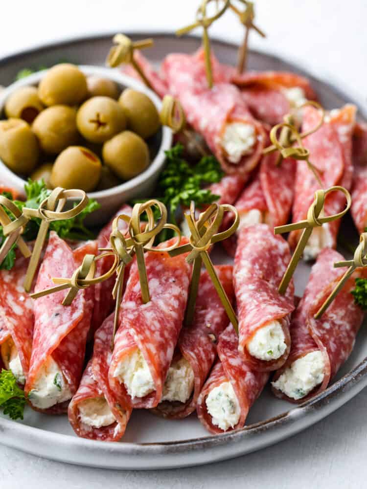 If you are looking for the perfect snack or appetizer, salami roll ups are your new best friend! Salami is wrapped around seasoned cream cheese for the perfect savory bite every time. Salami roll ups on a plate with a bowl of olives in the background.