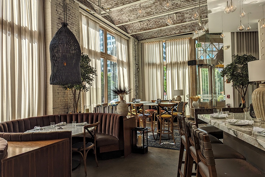 Empty restaurant interior during the day, featuring modern lightbulb light fixtures, long sheer-white curtains, and a half-circle brown banquette.