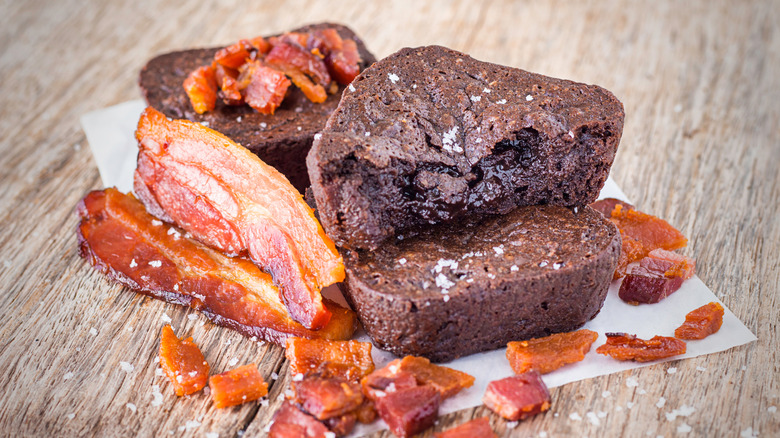 Two brownies in a stack with a bite taken out of the top one, and a third behind those covered with bacon bits and with slices of bacon to the side