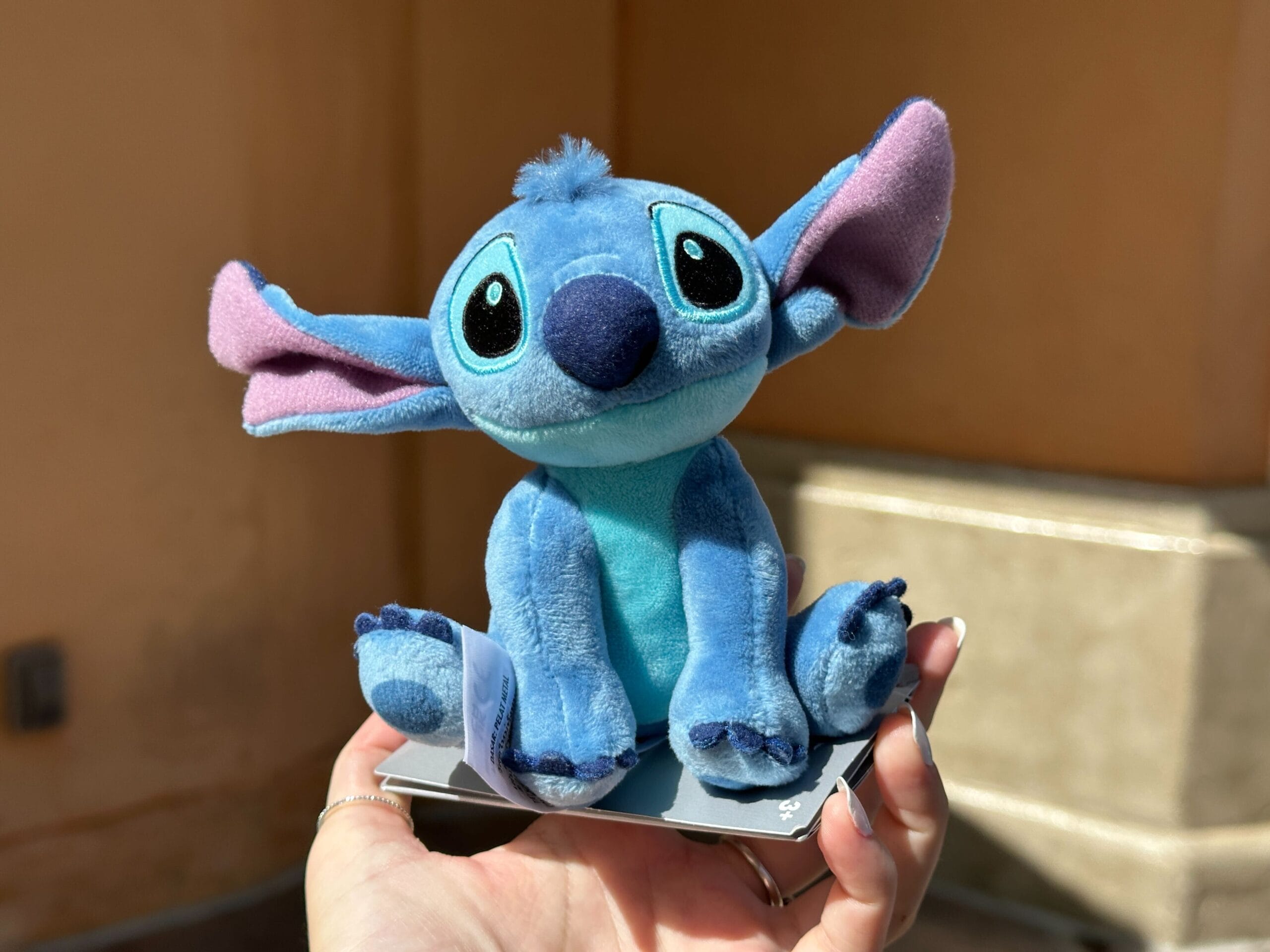 A hand holds a Stitch Shoulder Plush from Walt Disney World against a sunlit beige background.