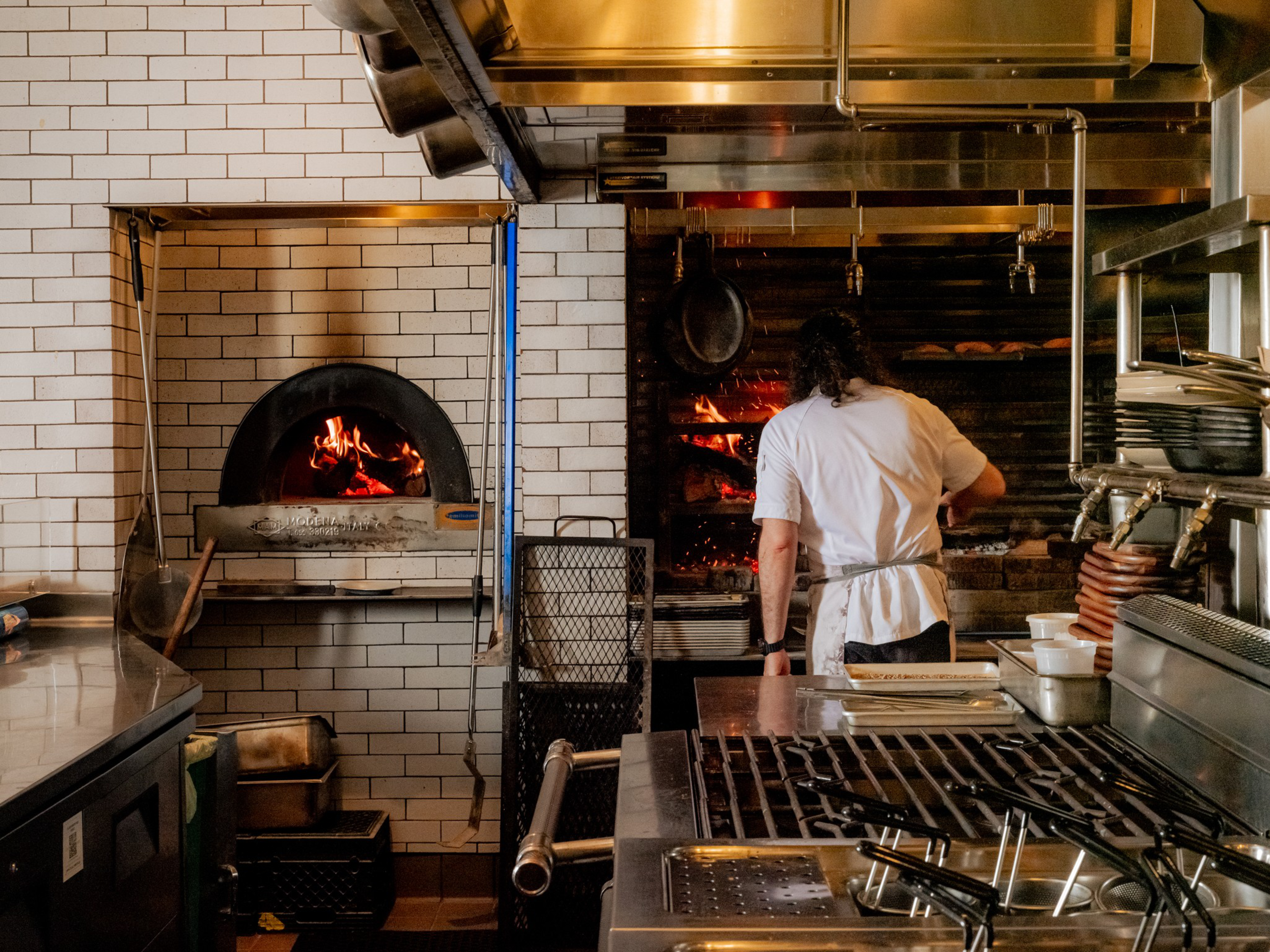 A chef in a white shirt tends to a wood-fired oven inside a kitchen with stainless steel appliances and white tiled walls.