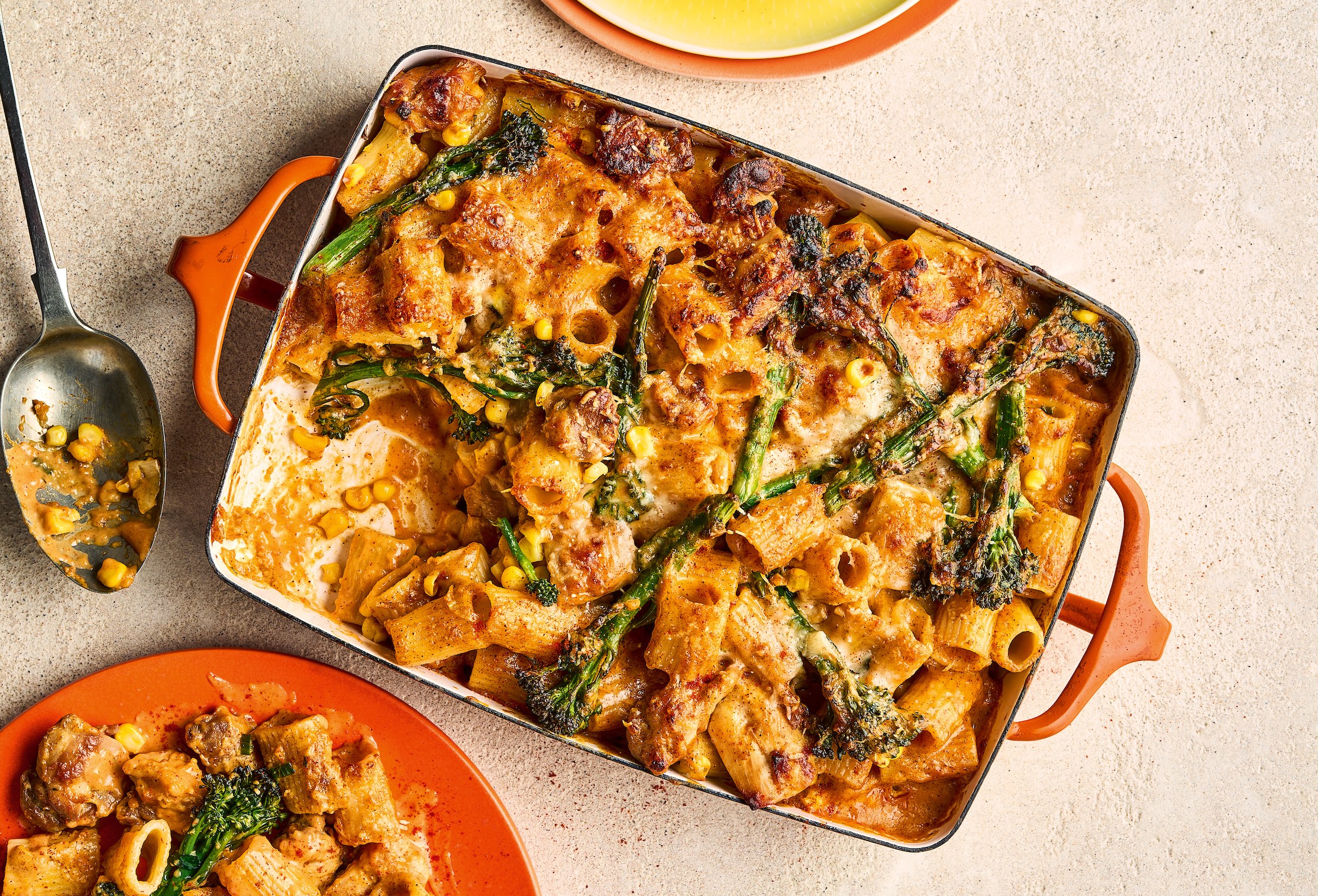 Smoky chicken pasta bake with broccoli and corn served in an orange baking dish and a small orange plate, with a large spoon.