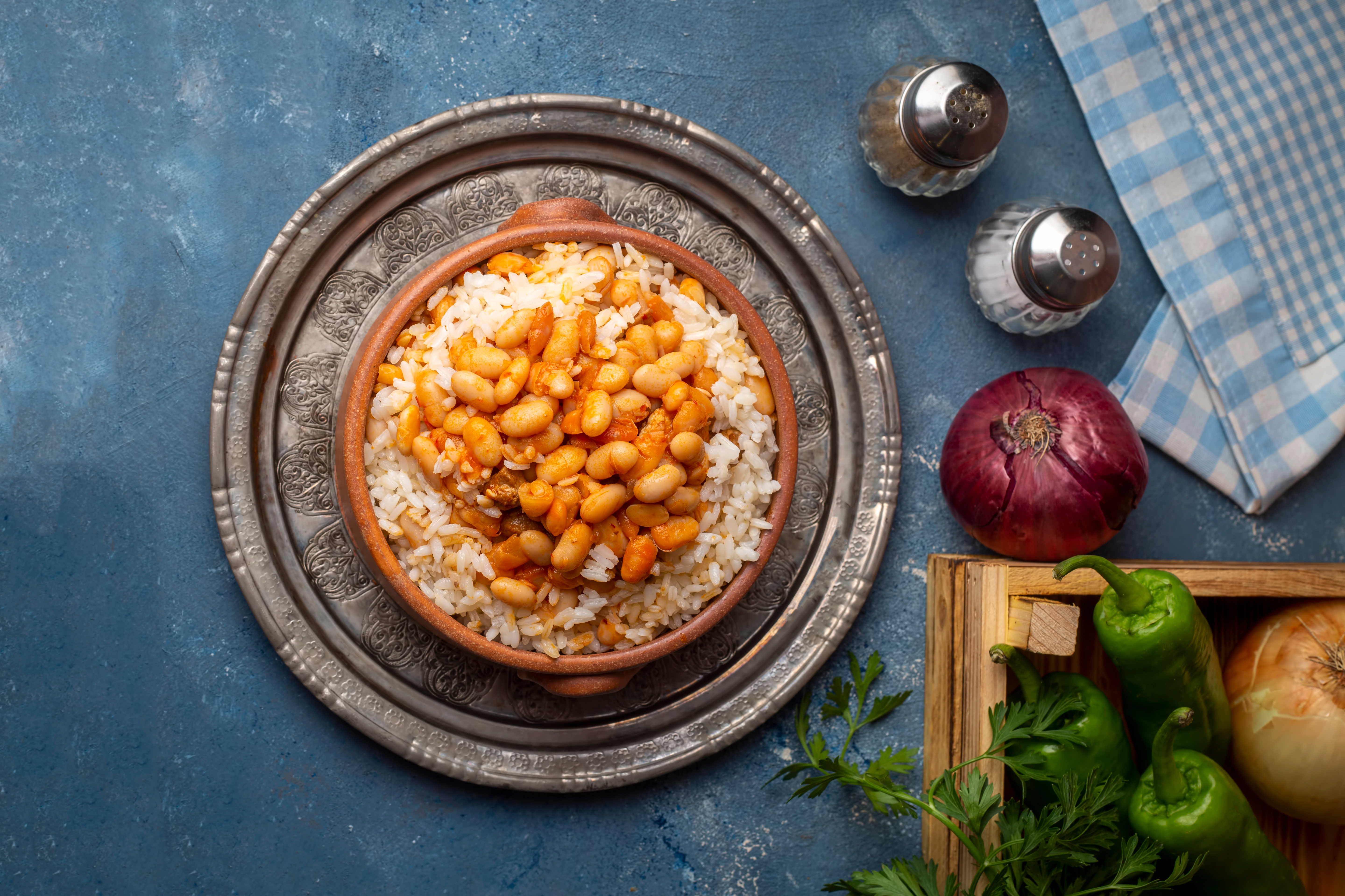 Kuru Fasulye with rice, Turkish haricot beans dish, on an ornate silver plate, next to an onion, peppers, parsley, and salt and pepper shakers.