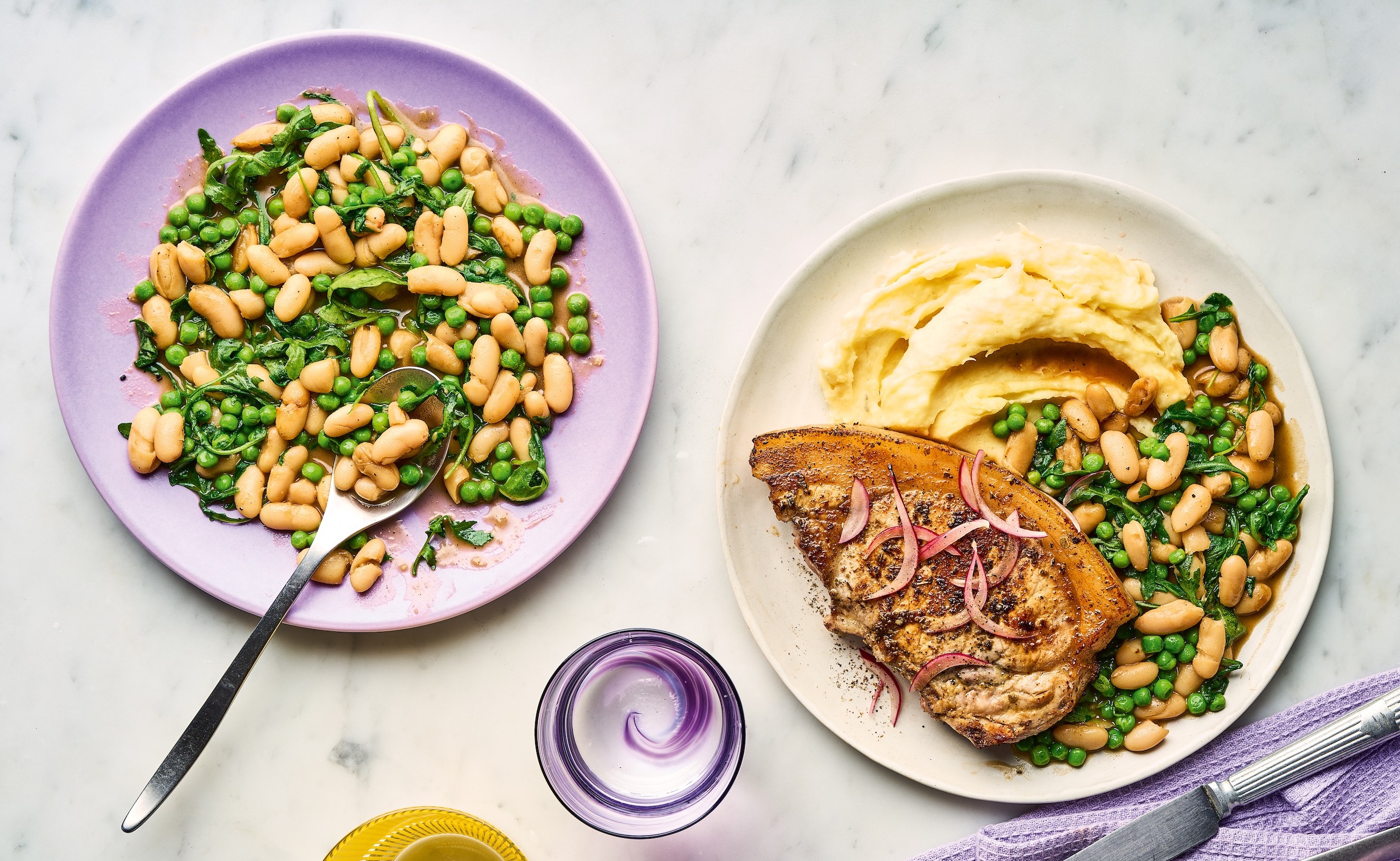 Plates of food on a white marble surface with a yellow glass and purple glass of water.
