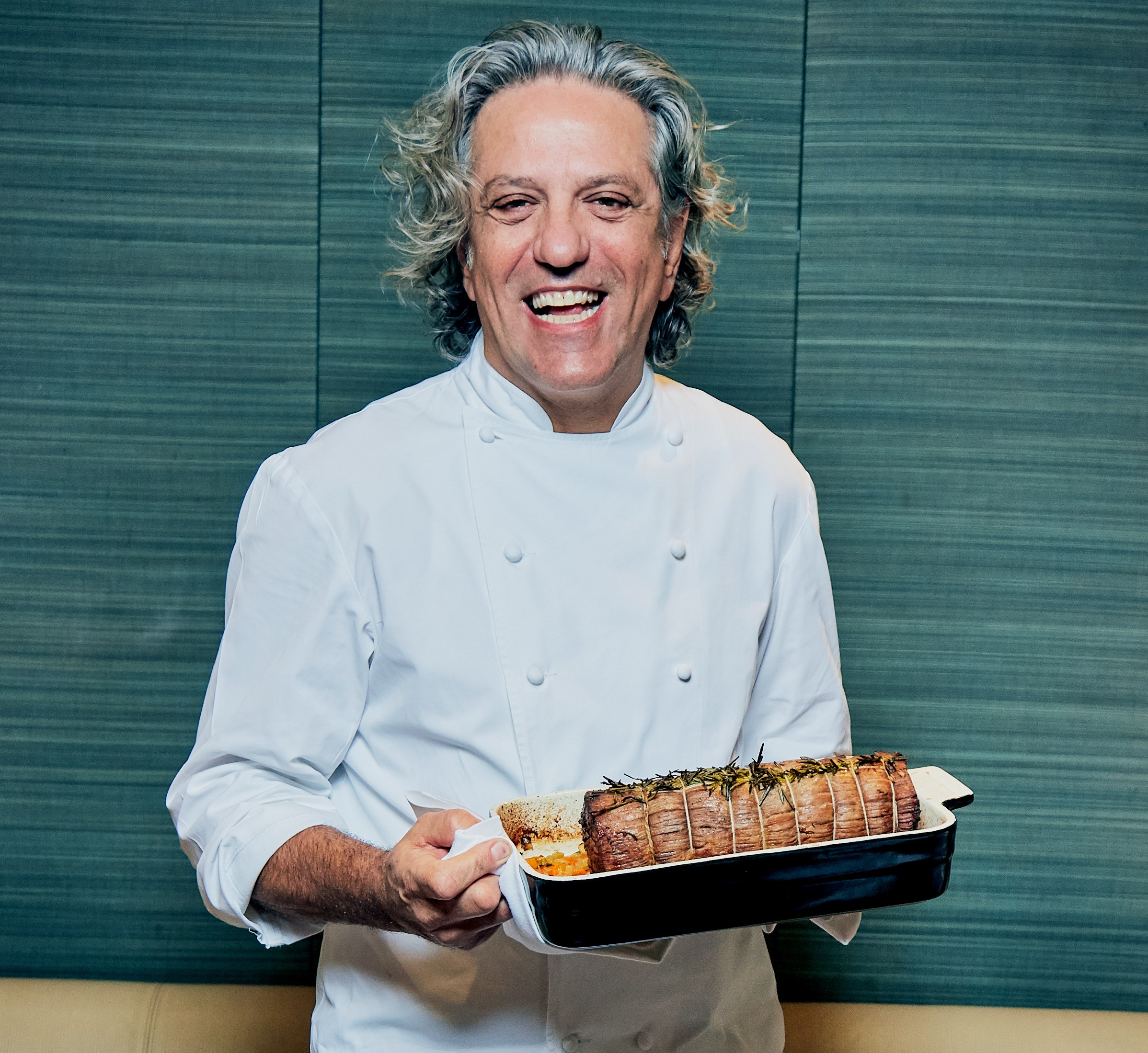 Giorgio Locatelli, a chef in a white uniform, smiles while holding a large roasted meat dish.