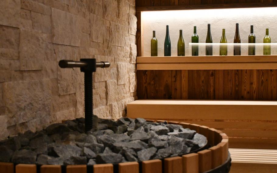 The interior of a sauna features light wooden benches with empty wine bottles as decor behind glass panels above the seating area. A central stove with stones on top has a T-shaped spout in the middle.