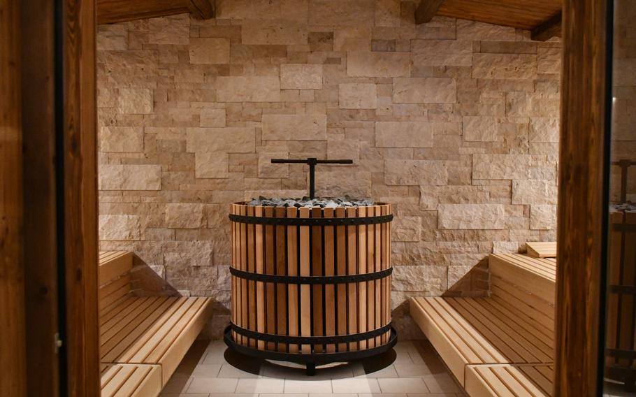 The interior of a sauna features light wooden benches on either side of a central wood-fired stove with stones on top with a T-shaped spigot in the middle. A tan stone tile wall provides the backdrop at the Südpfalz Therme.