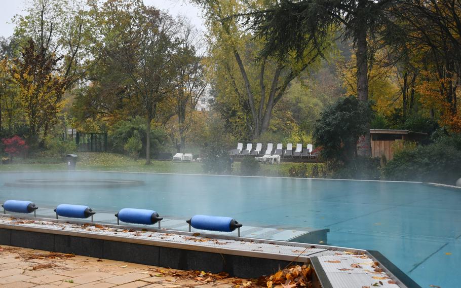 An outdoor swimming pool with steaming water is surrounded by trees, fall foliage is visible on a deck overlooking the pool.