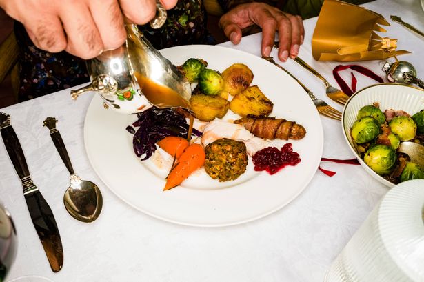 Man Pouring Gravy Over Turkey, Carrots, Potatoes, And Vegetables On Christmas Dinner Plate At Table