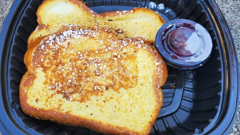 First Watch French toast with a tub of syrup on the side in a black plastic takeout container