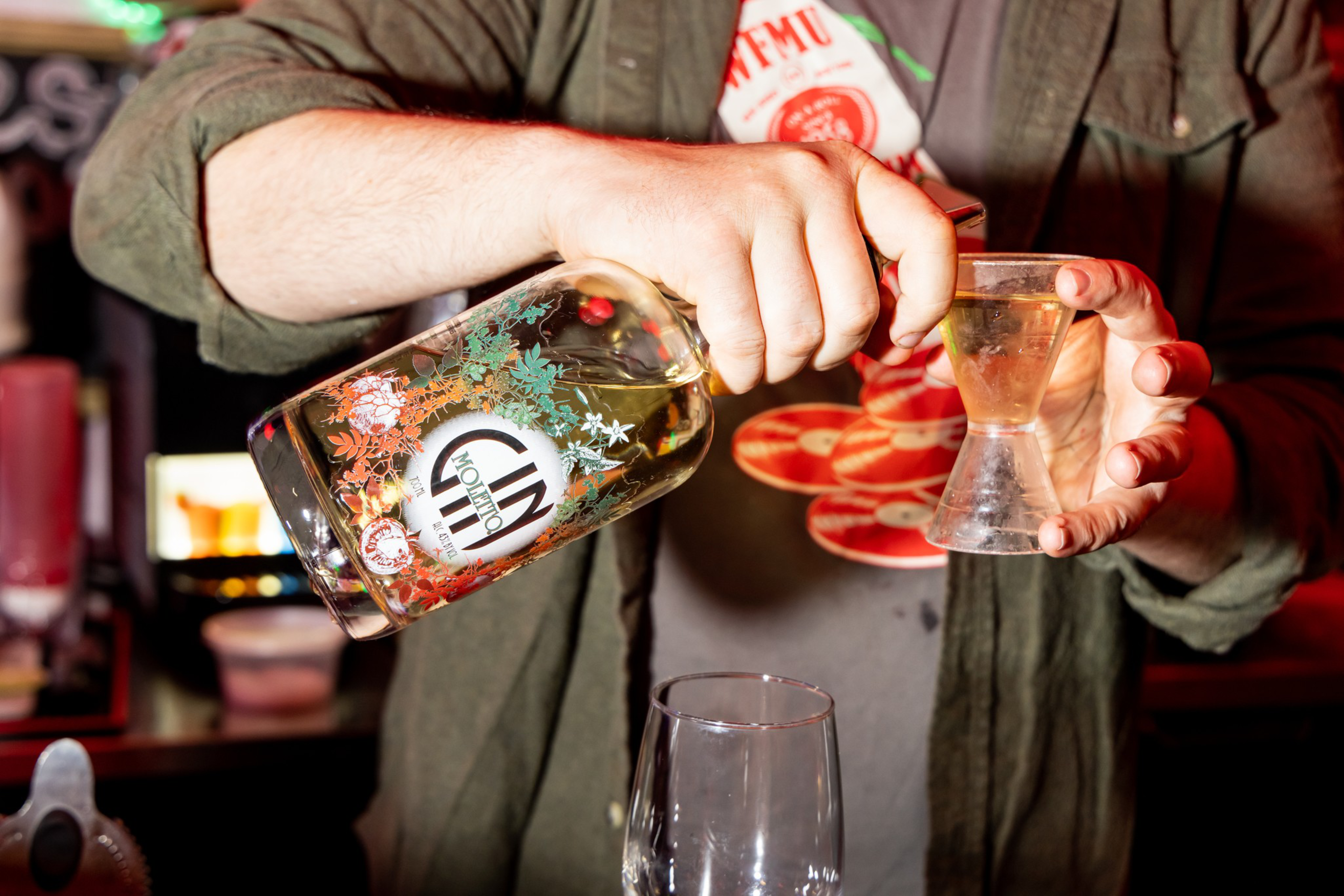 A person pours a clear liquid from a bottle labeled “Mojito” into a small measuring glass, with a larger empty glass below.