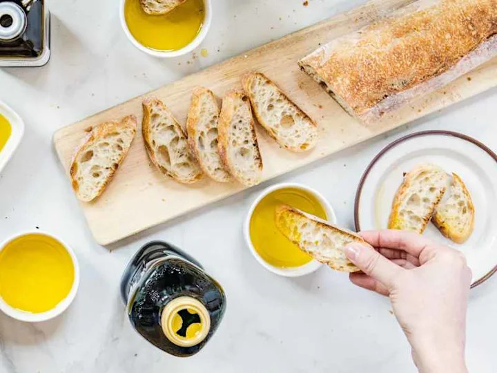 Olive oil bottles surrounded by ramekins of olive oil on table with sliced baguette on cutting board. Someone dipping bread into olive oil.