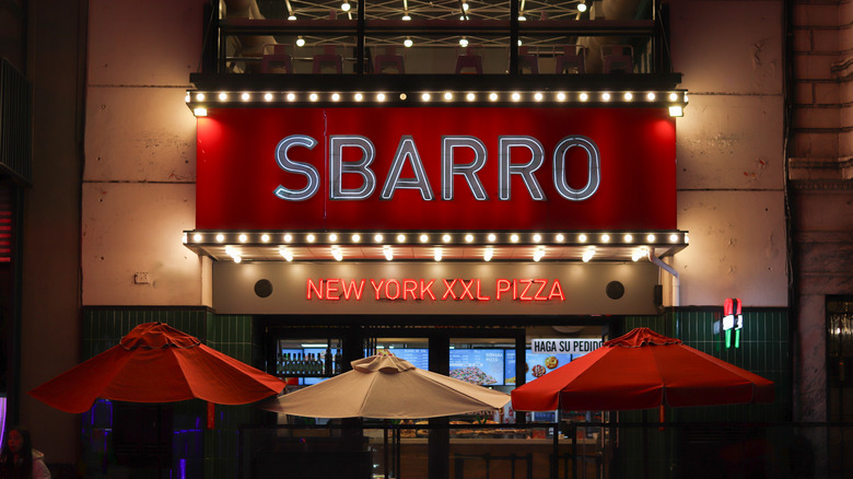 The illuminated sign on the outside of a Sbarro restaurant with tables and umbrellas outside