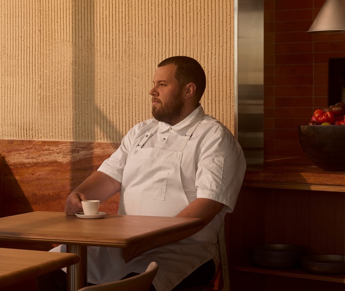 A man in chef's whites sits at a restaurant table with a coffee cup.