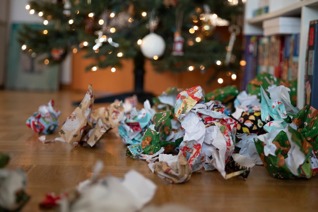 Discarded Christmas wrapping paper on a wooden floor near to a Christmas tree lit with fairy lights.