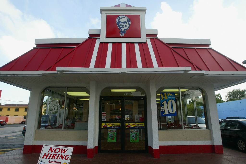 The exterior of a KFC restaurant with a red roof, a sign for Colonel Sanders, and a "Now Hiring" sign in the foreground.