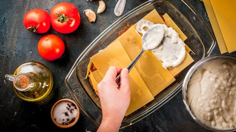 A home cook layers cheese sauce over lasagna with fresh tomatoes and seasonings
