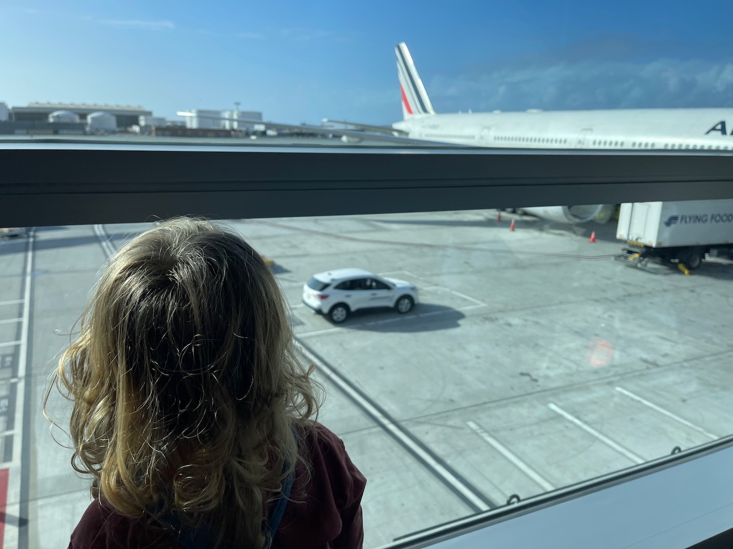 Child looking out airport window at Air France 777-300ER, observing aircraft and terminal activity.