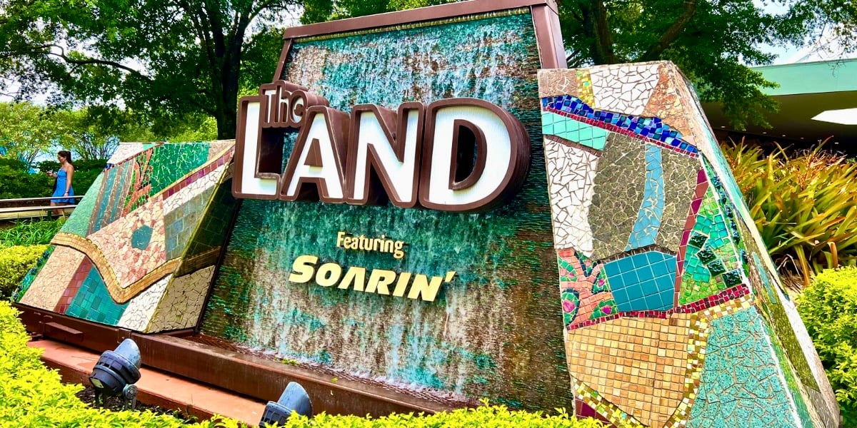 A colorful mosaic sign reads "The Land featuring Soarin'" with water flowing over it, surrounded by lush greenery and trees, near the entrance to Living with the Land.