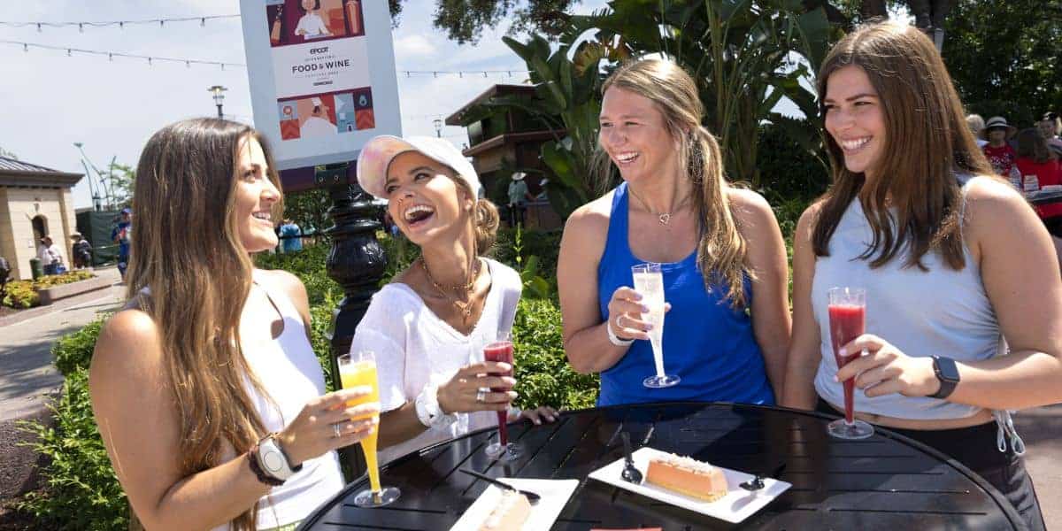 Four women are smiling and laughing while holding colorful drinks at an outdoor table with desserts, enjoying the sunny weather and greenery at the New EPCOT Lounge, with a food event sign in the background.