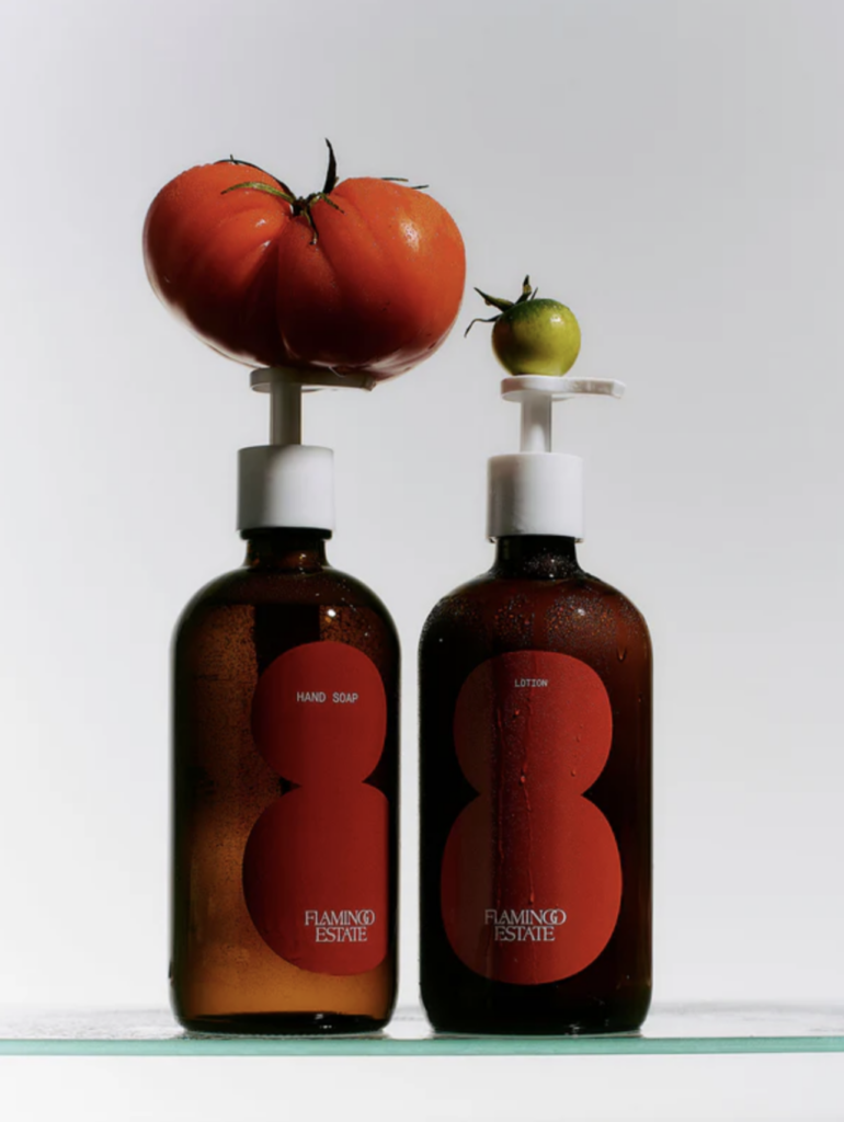 Two amber glass bottles with white pumps, each with tomatoes balanced on top of the pumps. The bottles have red labels that say Flamingo Estate.