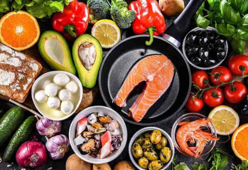 Assorted fresh foods including salmon, shrimp, avocado, vegetables, fruits, olives, bread, and cheese are arranged around a frying pan on a kitchen counter.