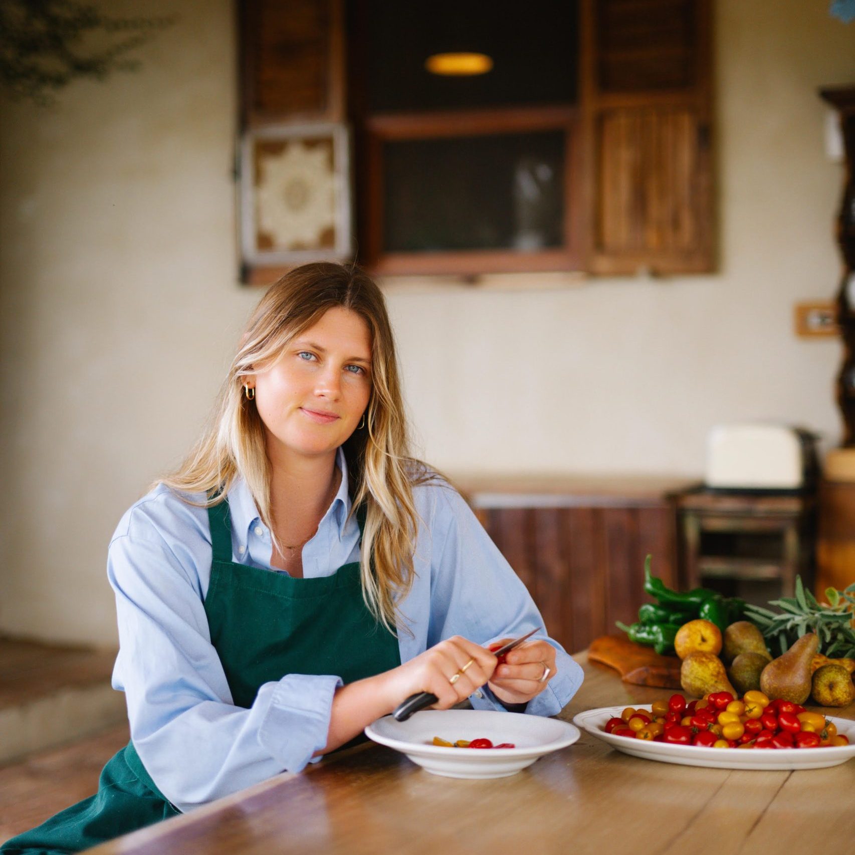 A woman with mid-length blonde hair wears a blue shirt and green apron. She sits at a table with fresh tomatoes and fruits.