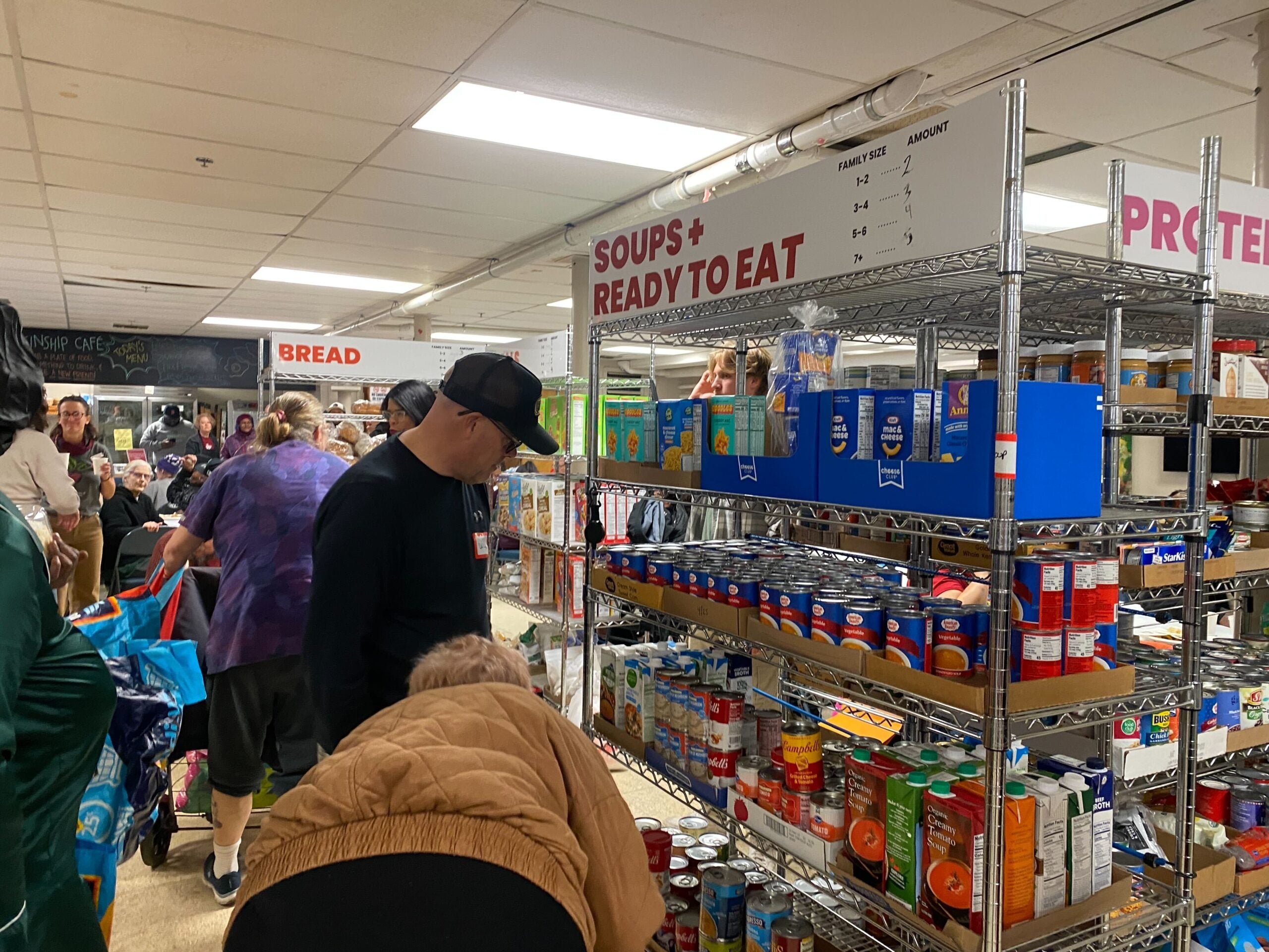 People browse shelves stocked with canned goods and soups in a crowded food pantry. Signs above the shelves label sections such as Soups + Ready to Eat and Bread.