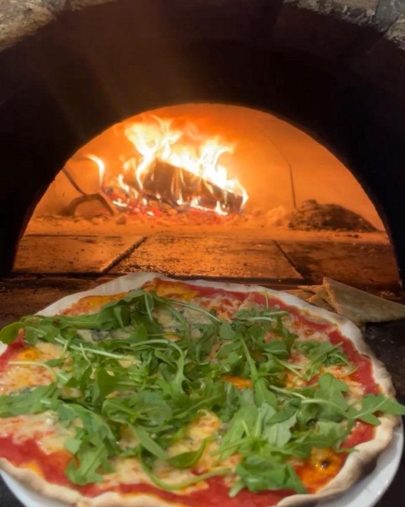 a La Gita Pizza covered in greens sitting outside a wood-fire oven