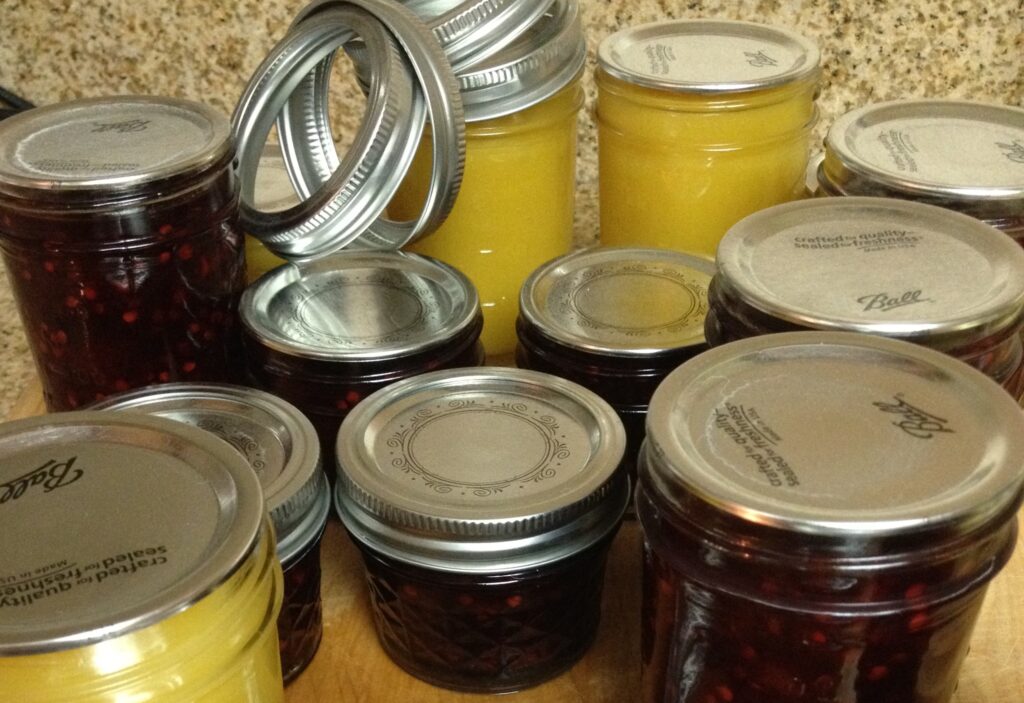 Assorted clear class canning jars, some filled with creamy yellow curd and others with dark red jam, sit on a cutting board with the rings close by to screw on top of the lids.