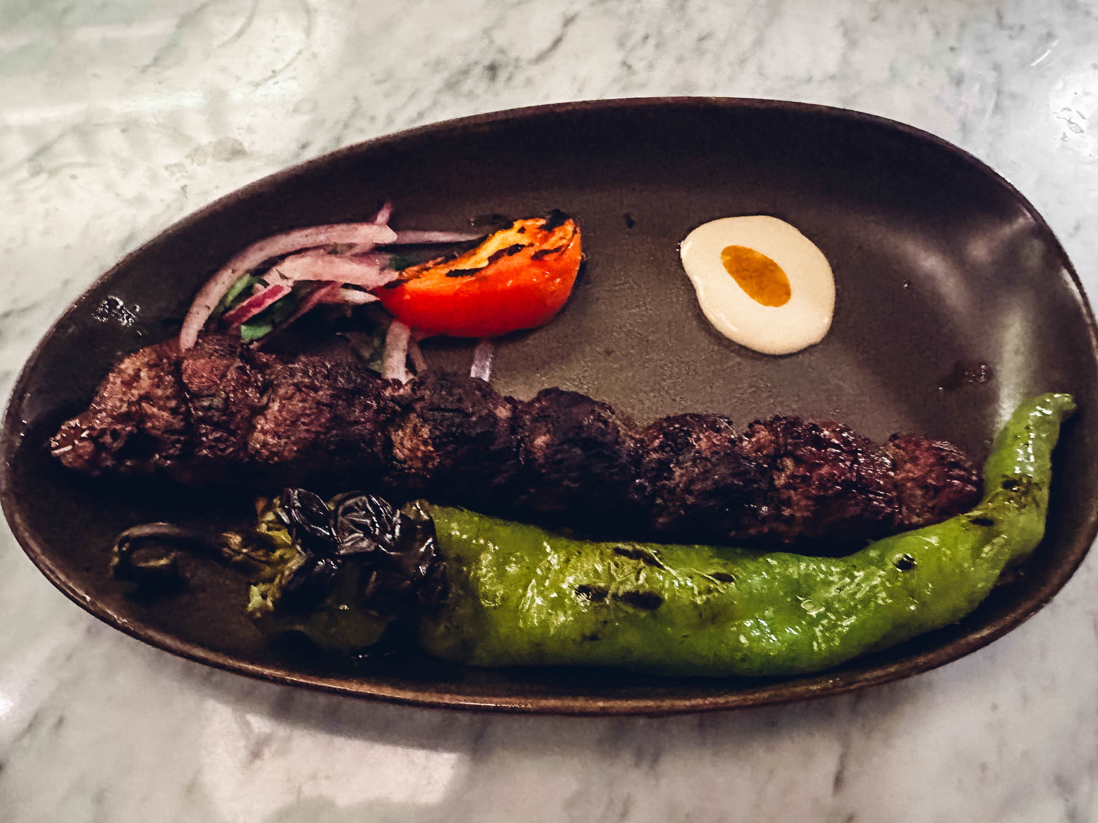 A kebab on a plate next to a long pepper.