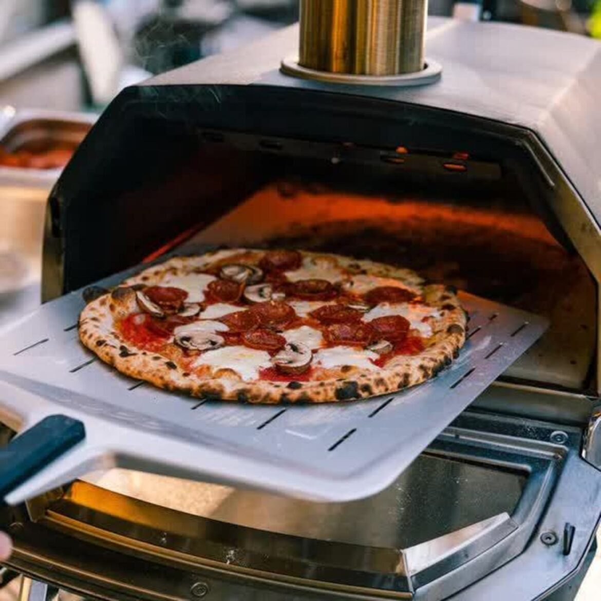 The Best Gifts for Cooks, Chefs & Foodies in 2025 A small metal pizza oven with the door open and a pepperoni and mushroom pizza being pulled out of the oven with a pizza peel.