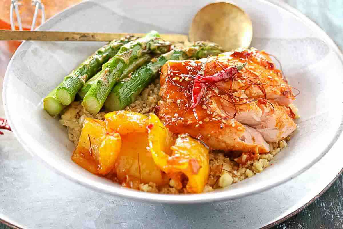 A white bowl with grilled chicken, asparagus, roasted yellow bell peppers, and quinoa, garnished with sesame seeds and chili threads, with a spoon on the side.