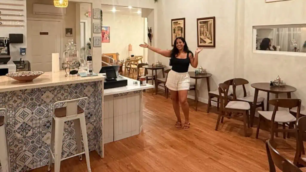inside a coffee shop and wine bar with its owner posing at the center. She's wearing white shorts and a black t-shirt. Small tables are set against the wall making an L shape around a bar.
