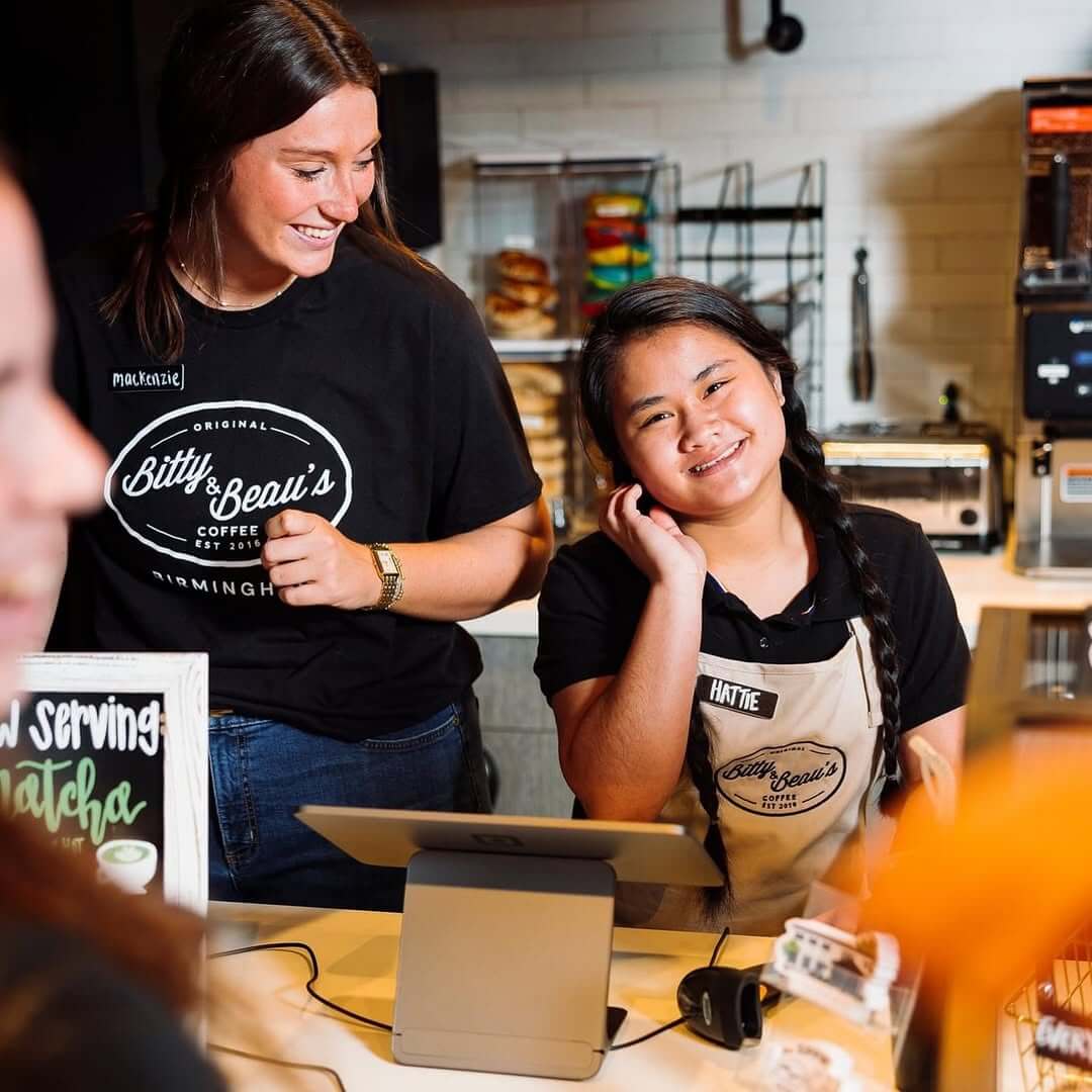 7 Beloved BHAM Restaurants We'll Miss - 7 Two women stand behind a coffee shop counter; one wears a black T-shirt and smiles, while the other, wearing an apron and name tag, faces the camera with a hand near her ear at Bitty and Beau's Cafe in Homewood.