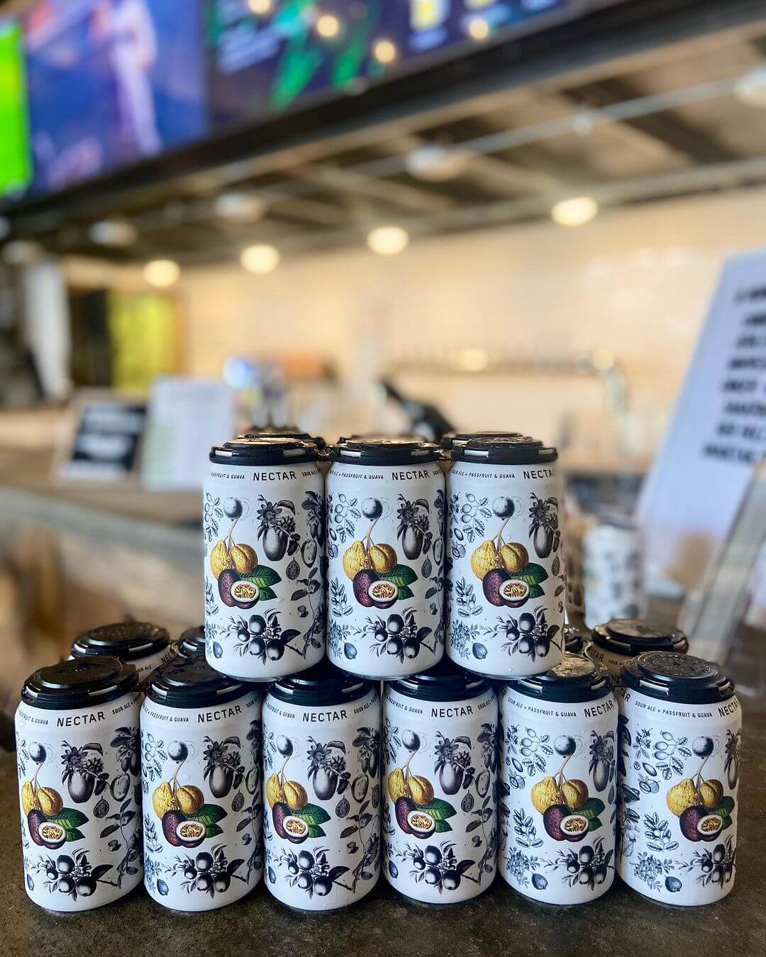 7 Beloved BHAM Restaurants We'll Miss - 6 A stack of twelve white aluminum cans with fruit illustrations and the word "NECTAR" printed on them, displayed on a counter at Uproot Brewing.