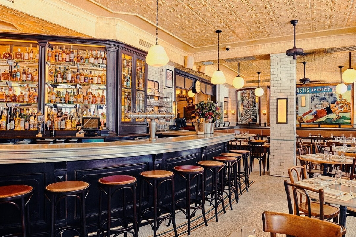 A well-lit Paris-inspired restaurant interior with a long bar, high stools, round hanging lights, and shelves stocked with bottles, surrounded by neatly set dining tables—perfect for a stylish day trip in the heart of Nashville.