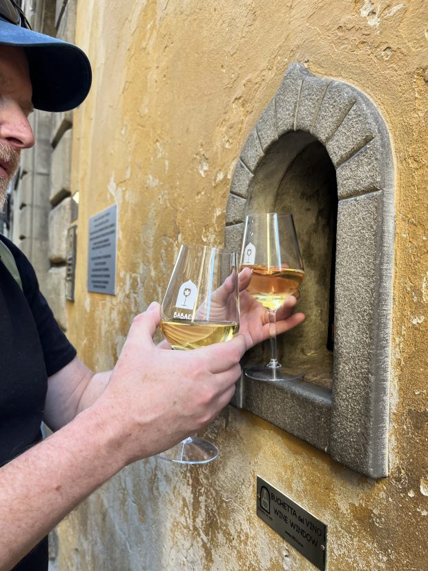A man grabs two glasses of wine that are being served out of a wine-sized hole in the wall