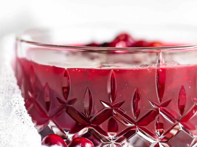 A close-up of a crystal bowl filled with red cranberry sauce, with whole cranberries visible inside and around the bowl.