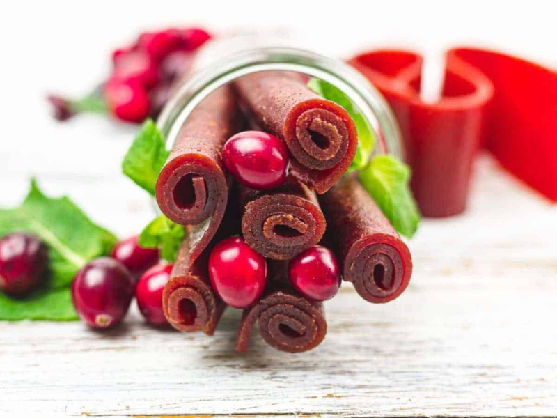 Rolled cranberry fruit leather strips and fresh cranberries are arranged on a white wooden surface, with a glass jar and green leaves in the background.