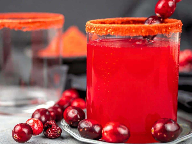 A glass of bright red drink with a chili powder rim, garnished with cranberries, sits on a table with fresh cranberries scattered around. Another glass is in the background.