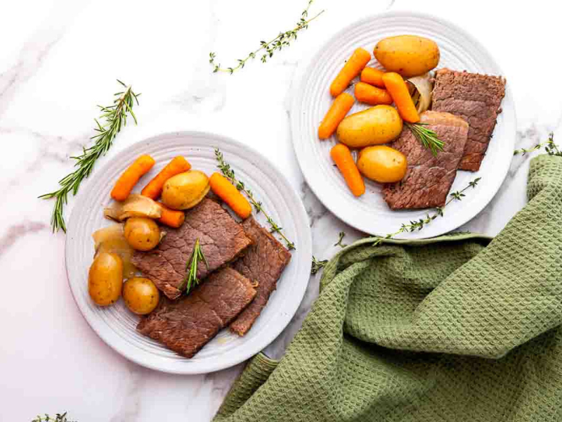 Two white plates with slices of roast beef, potatoes, carrots, and onions, garnished with rosemary, on a white surface with a green textured cloth.