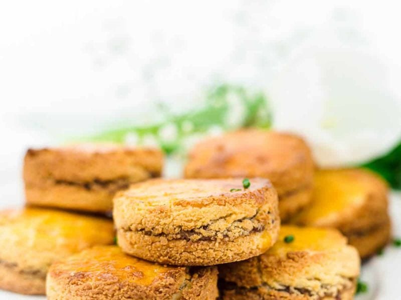 A close-up of several stacked golden-brown biscuits with a visible filling, set against a blurred white background with a hint of greenery.