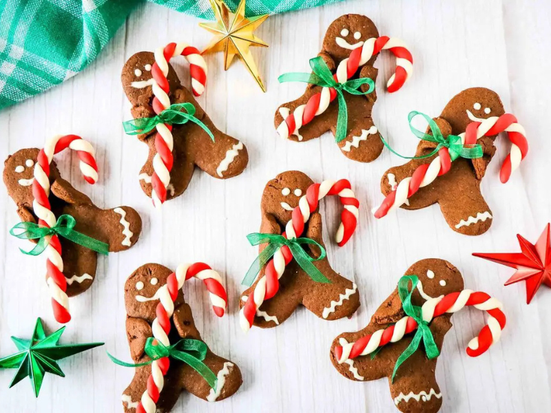 Gingerbread cookies shaped like people, each holding a small candy cane tied with a green ribbon, are arranged on a white surface with colorful star ornaments nearby.