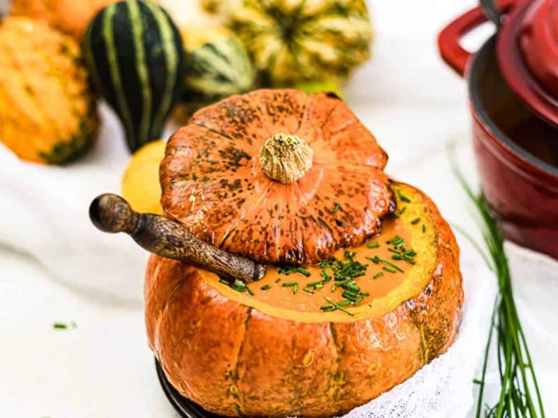 Pumpkin soup served in a hollowed-out pumpkin with its lid resting on the side, garnished with chopped chives and a spoon. Other gourds and a red pot are in the background.
