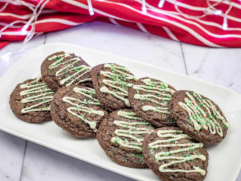 A white plate with nine chocolate cookies drizzled with white icing and green and red sprinkles, set on a marble surface with a red striped cloth and holiday string lights in the background.