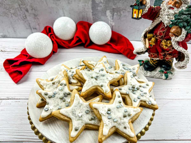 Plate of star-shaped cookies with white icing and silver sprinkles, next to a Santa figurine, white ornaments, and a red cloth on a white wooden surface.