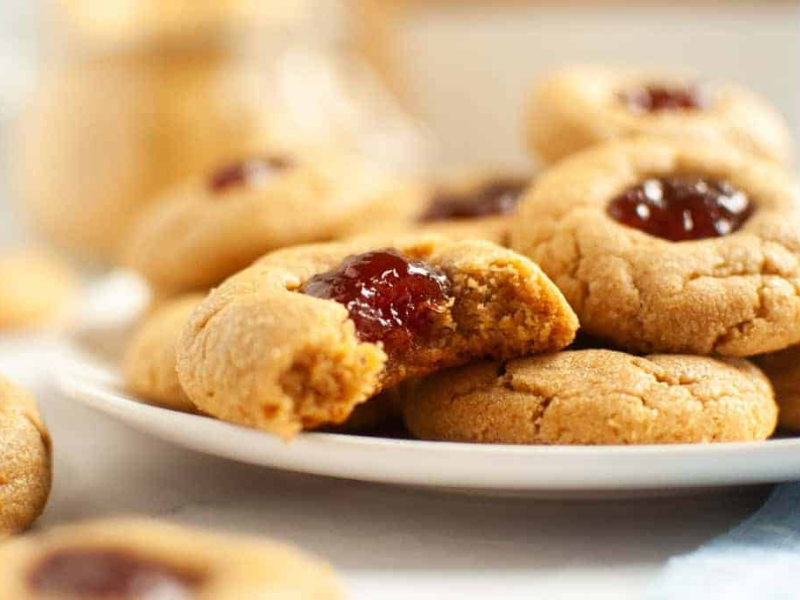 A plate of peanut butter thumbprint cookies filled with red jelly; one cookie is partially eaten, showing the inside.